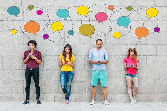 Four people leaning against a grey wall with colored speech bubbles, using their smartphones.