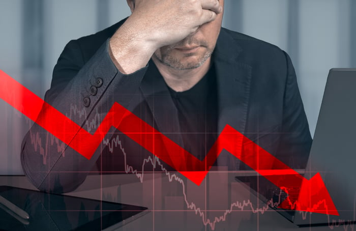 A person with their head in their hands and a down arrow on an overlay of a stock graph.