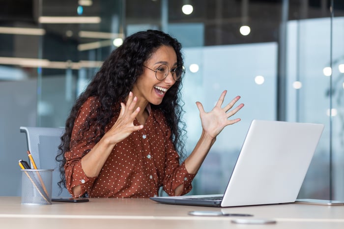 A person waves their hands with excitement while sitting at a table and looking at a computer.