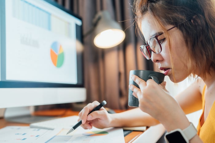 Investor at a desk sipping  from a mug and looking at financial charts on papers and a computer monitor.