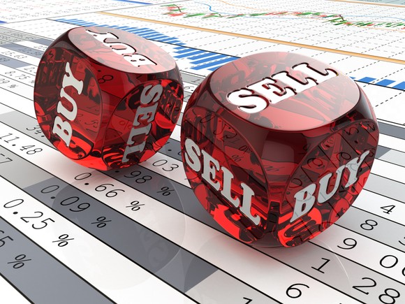 Two red dice that say buy and sell being rolled across printouts displaying financial data.