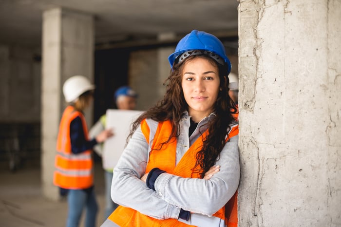 A construction worker leaning against a wall with people in the background looking at blueprints.