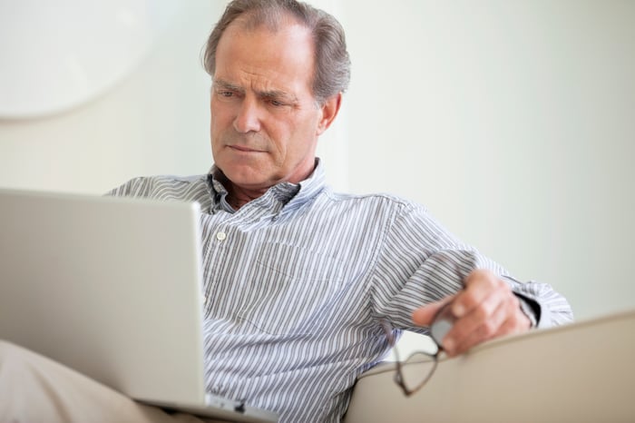 A person sitting on a couch and holding their glasses while closely reading content from an open laptop.
