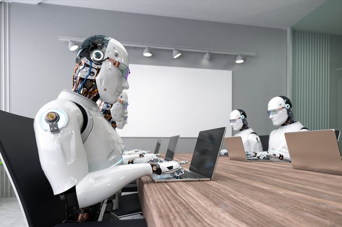 Artificial intelligence powered robots sitting at a conference room table typing on laptops.