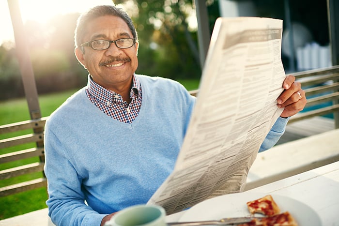 Person sitting on bench outdoors, holding a newspaper and smiling. 