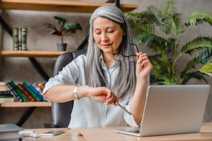 Someone is sitting in front of an open laptop, looking at their watch.