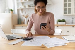 Smiling person looking at smartphone and typing on calculator