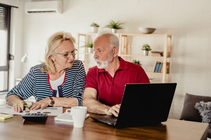 A senior couple at a table with a laptop open, calculator in front of them, and various papers scattered.