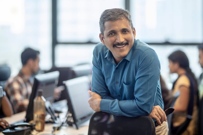 Person in office, leaning on the back of a chair and smiling.