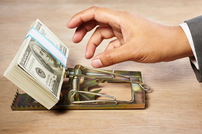 A hand reaching for a neat stack of $100 bills in a mouse trap.