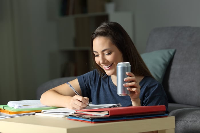 A smiling person writing in a notebook drinking an energy drink from a can.