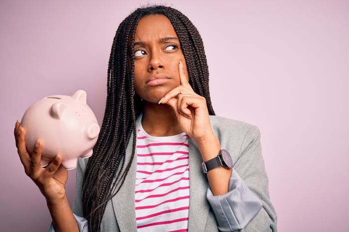 A person with a thinking or questioning expression on their face is holding a piggy bank.