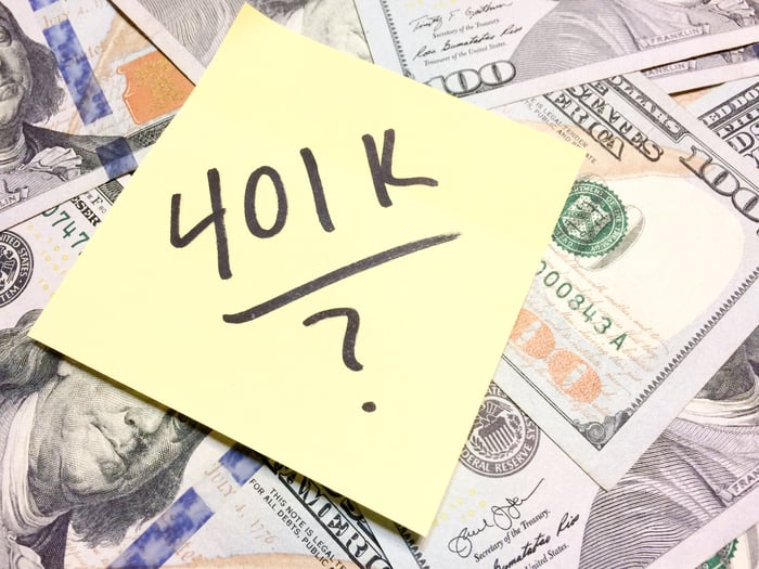A post-it note with 401k and a question mark written on it laying on top of a pile of hundred-dollar bills.