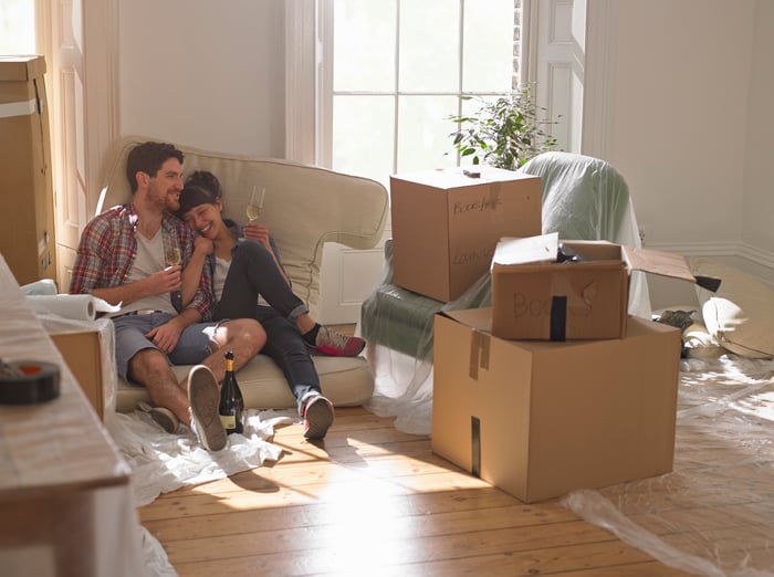 A smiling couple surrounded by boxes, sitting on a couch cushion on the floor of the new home they're moving into.