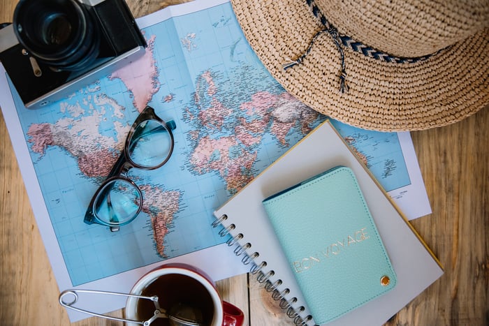A map laid out on a table with a camera, notebook, coffee cup, hat and travel itinerary.