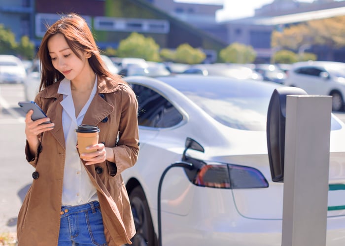 A person on their phone stands in front of their car that is plugged in at a charging station.