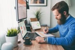 bearded person at desk by computer taking notes