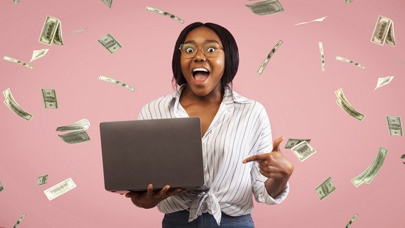 A happy person is showered with cash while checking a laptop.