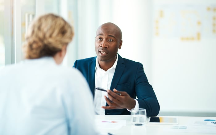 A person gestures with a pen while reviewing documents on a table across from another person.