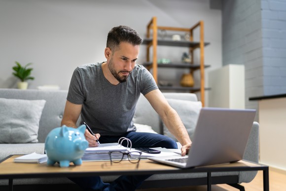 Someone writing and looking at a laptop with a piggy bank on the table.