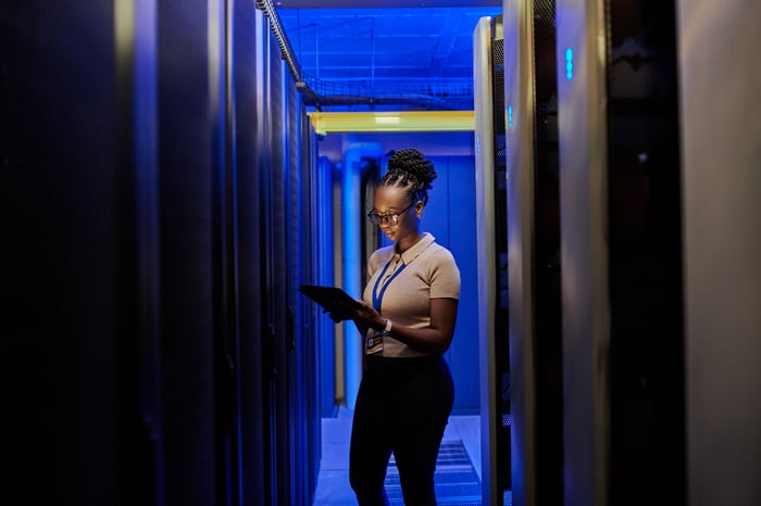 A person standing in a dark server room looking down at a tablet device.