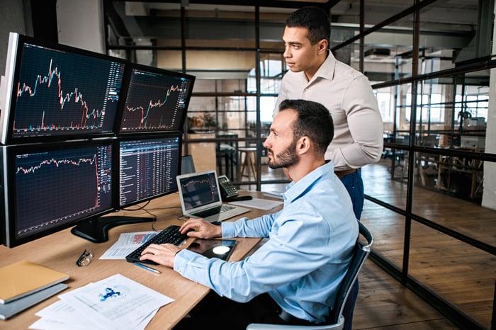 Two investors looking at a series of computer screens with price charts on them.