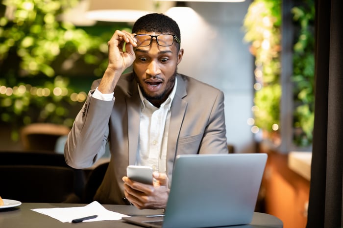 A business person looking at their phone with an astonished look while sitting in front of their laptop.