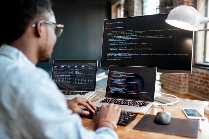 A person sitting at a desk coding on two laptops and a monitor.  