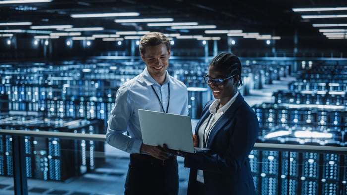 Two smiling employees looking at a laptop computer inside a data center.