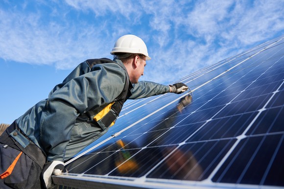 A person wearing a hard hat and other personal protective equipment works on a solar panel with a bright blue sky in the background.