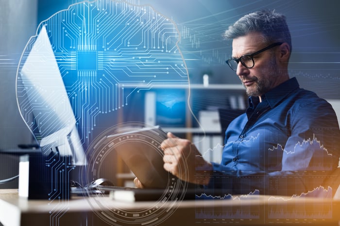 A person looking at a mobile device while seated at a computer desk with an overlay of AI algorithms and stock price graphs.