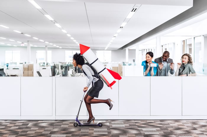 Person riding through cubicle farm on scooter with a rocket strapped to their back.