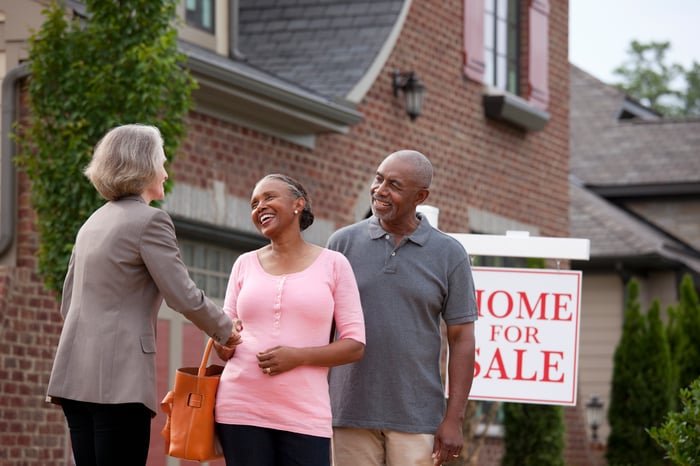 A real estate agent standing with their clien'ts outside their home, preparing to list it for sale.