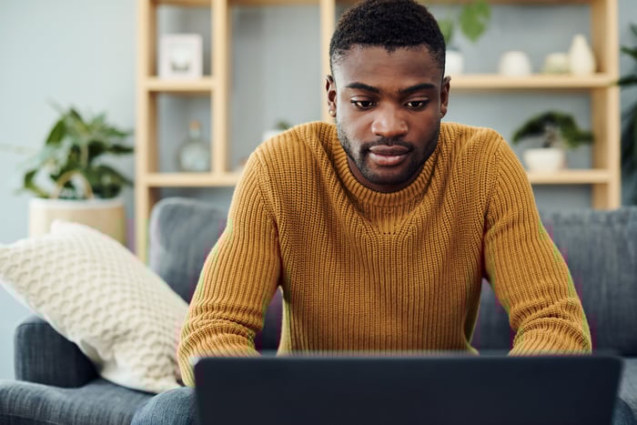 A person seated on a couch in their home who's typing on a laptop.