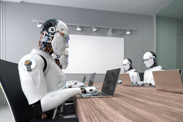 Multiple humanoid robots typing on laptops while seated at a table in a conference room.