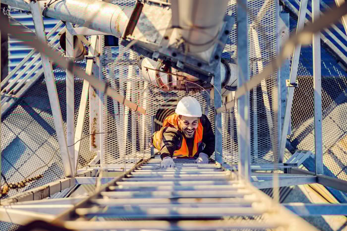 A person wearing personal protective equipment smiles while looking up and climbing a ladder.