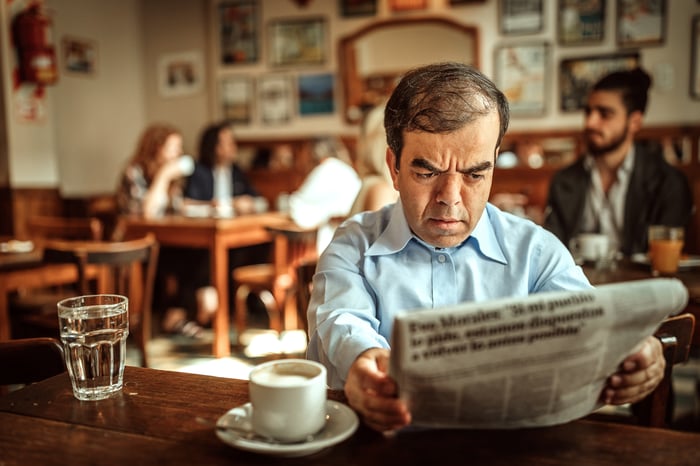 Someone is seated by a cup of coffee, reading a newspaper and looking worried.