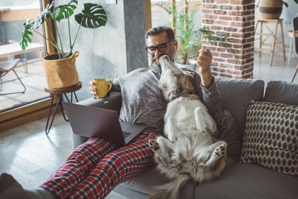 person sits on couch with dog, coffee, and computer