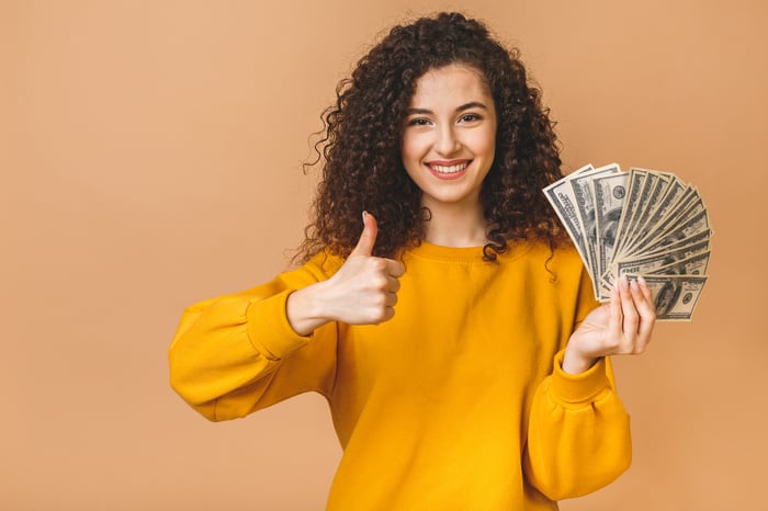 A person holding a fan of money while making a thumbs-up sign.