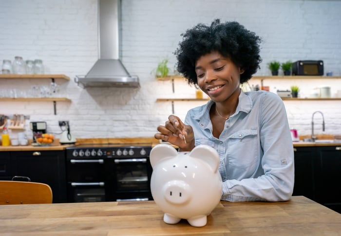 A person smiling while leaning on their kitchen counter and dropping money into a piggy bank.