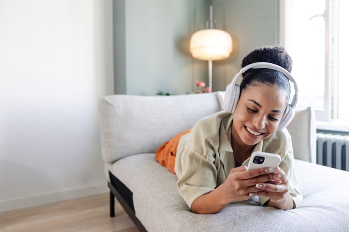 A smiling person laying on the couch with headphones on while looking at a smartphone.