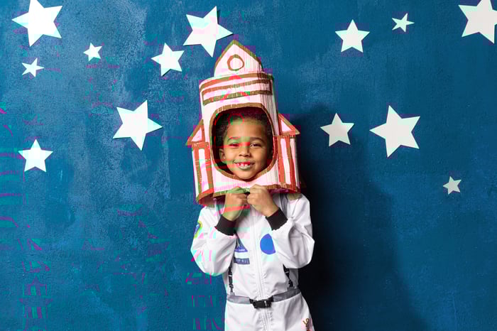 A child in a spacesuit with a handcrafted rocket on their head.