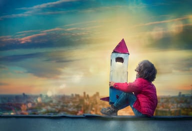 Small child hugging a wooden toy rocket while sitting on a rooftop looking wistfully at the sky.