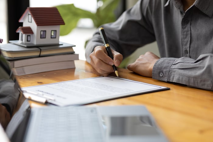 A person signs paperwork with a miniature model home in the background.