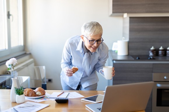 A woman stands in her kitchen holding a coffee cup while looking at her laptop sitting on a table with a smile on her face