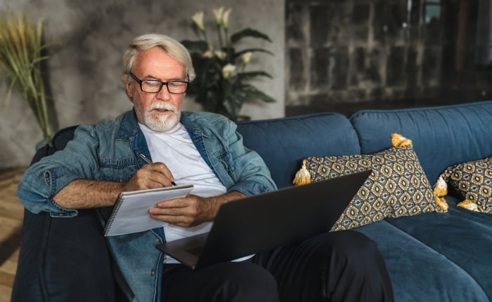 Serious person with laptop on lap writing in notebook.