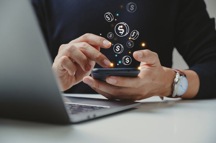 A person with a laptop looking at a mobile device that has images of coins coming out of the screen.