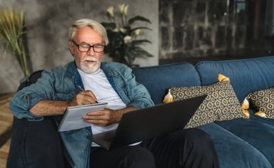 Senior holding laptop and writing note in notebook