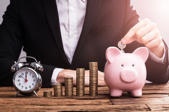 A person dropping a coin into a piggy bank, beside which is an alarm clock and ascending stacks of coins.