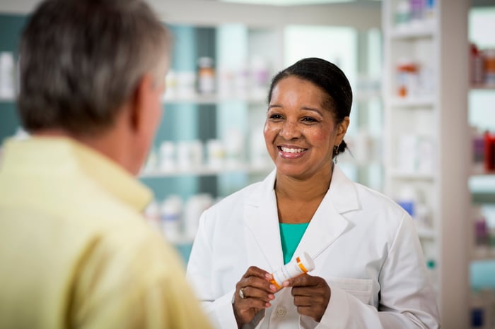 A smiling pharmacist holding a prescription bottle while speaking with a customer.